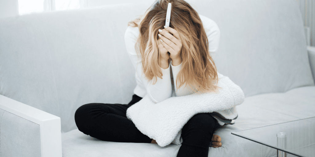 Young woman in black pants and white shirt sets on a white couch holding a white blanket. She is holding a pregnancy test with both hands and has her arms covering her face, appearing to be frustrated