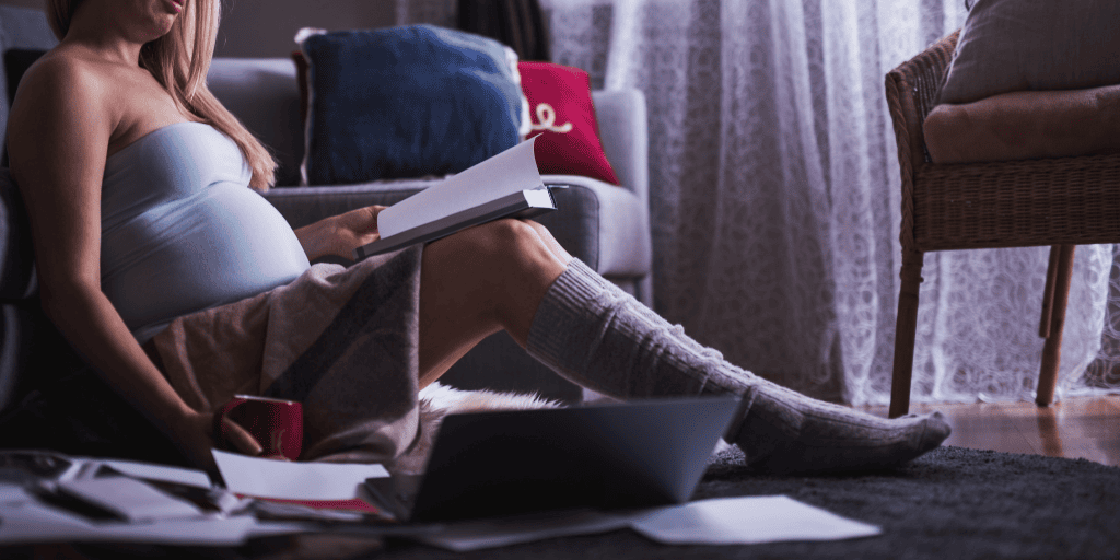 Pregnant student studies at home while setting on the floor surrounded by books, her computer, and a coffee mug